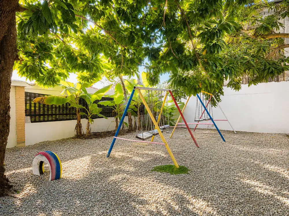 Swings and a painted tyre in the gravel play yard under a mango tree